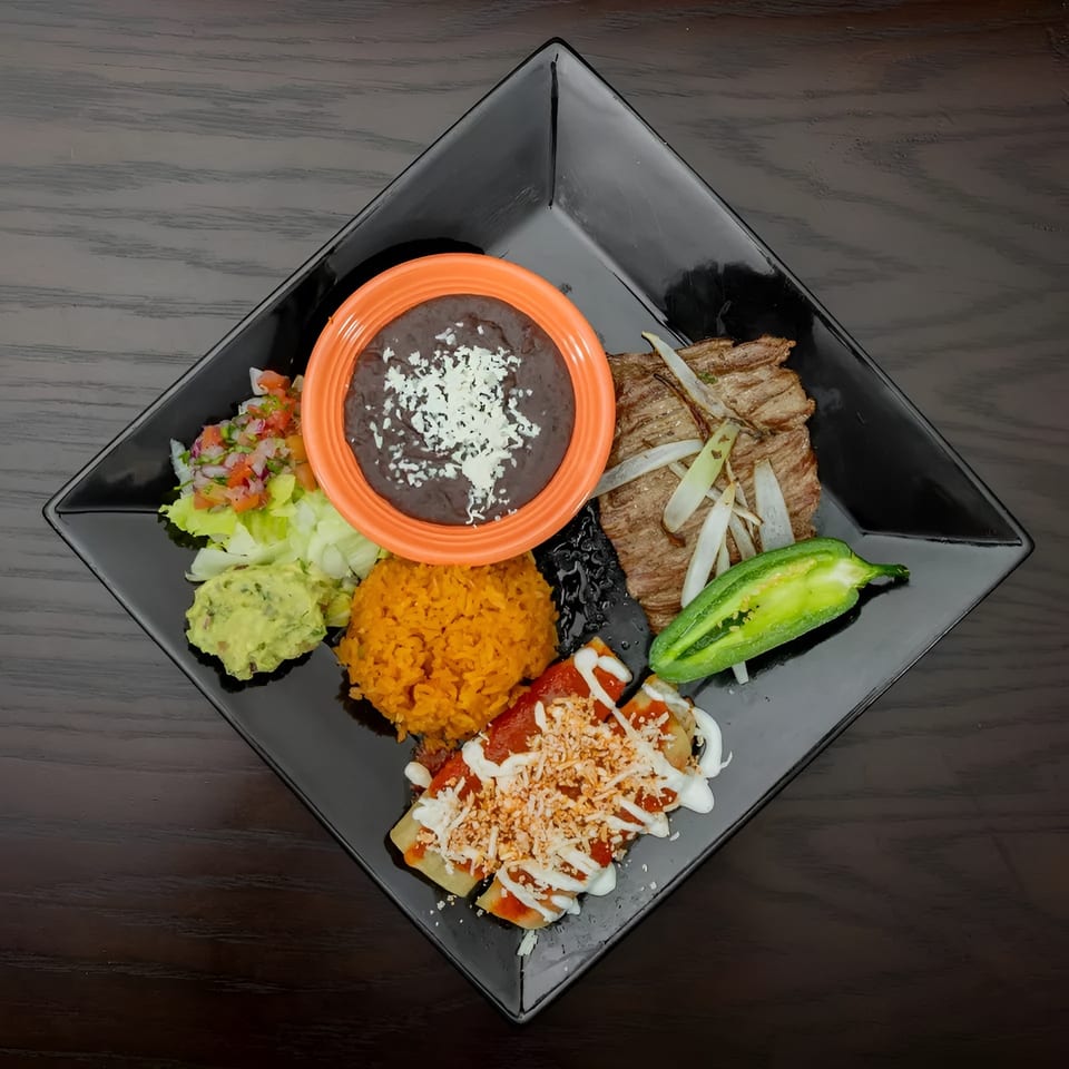 A black square plate holds a Mexican combo meal with steak, enchiladas, rice, beans, guacamole, and salsa.