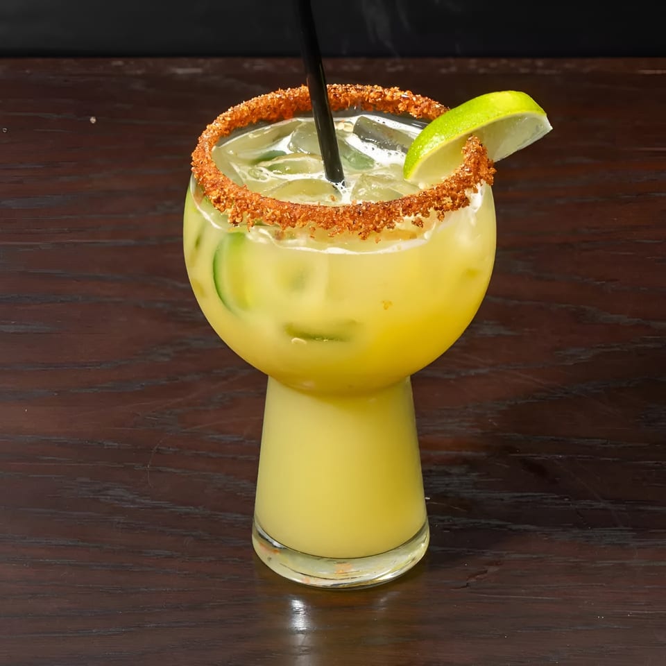 A yellow cocktail with cucumber slices, ice, a lime wedge, and a chili-salted rim on a wooden table.