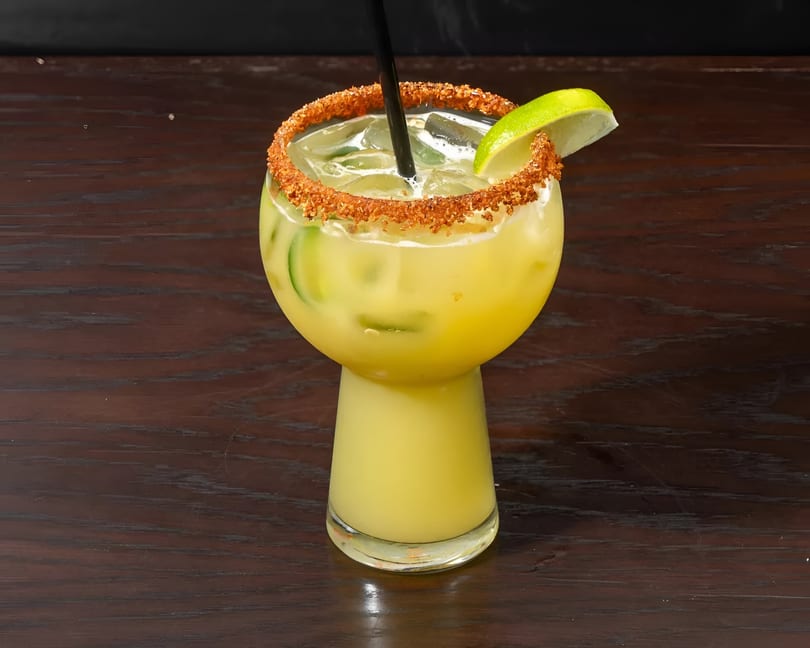 A yellow cocktail with cucumber slices, ice, a lime wedge, and a chili-salted rim on a wooden table.