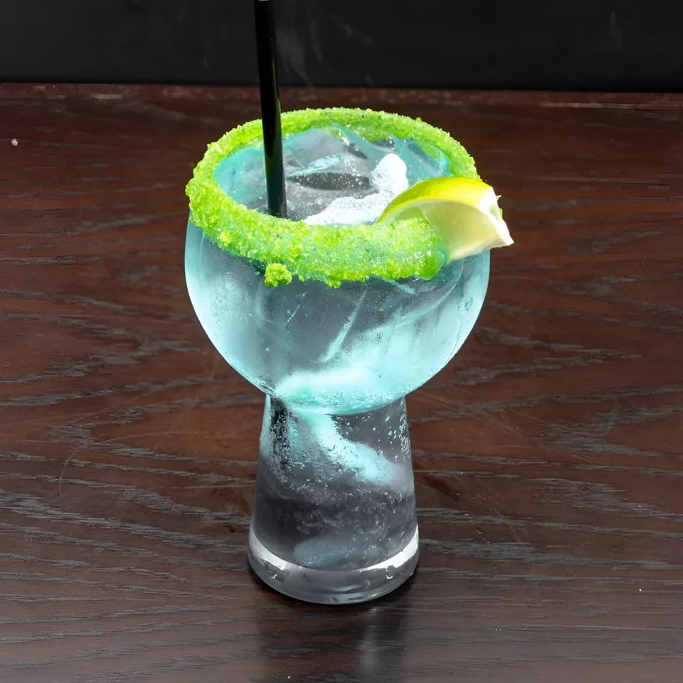 A vibrant blue cocktail with a bright green sugar rim and a lime wedge on a dark wooden table.