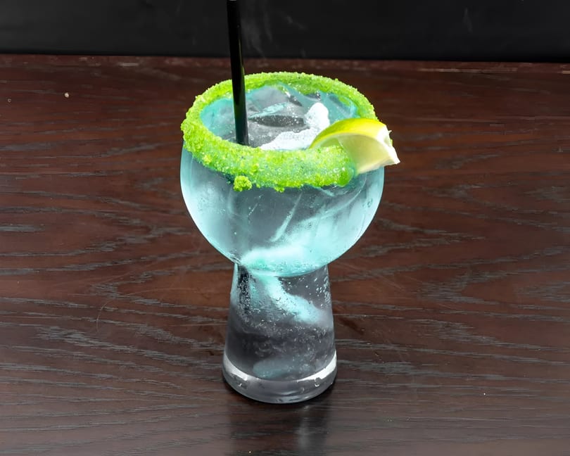 A vibrant blue cocktail with a bright green sugar rim and a lime wedge on a dark wooden table.