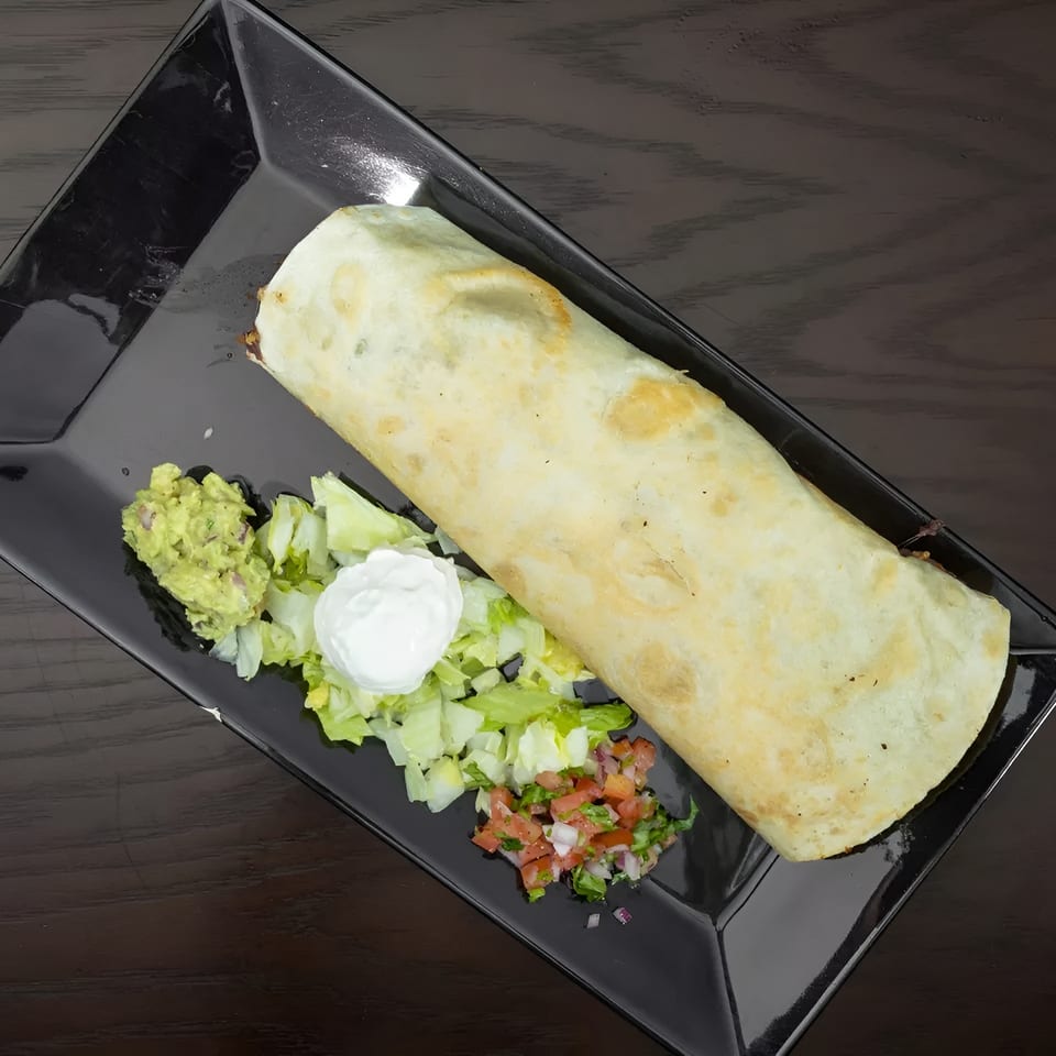 Large burrito on a black plate with sides of guacamole, shredded lettuce, sour cream, and pico de gallo.