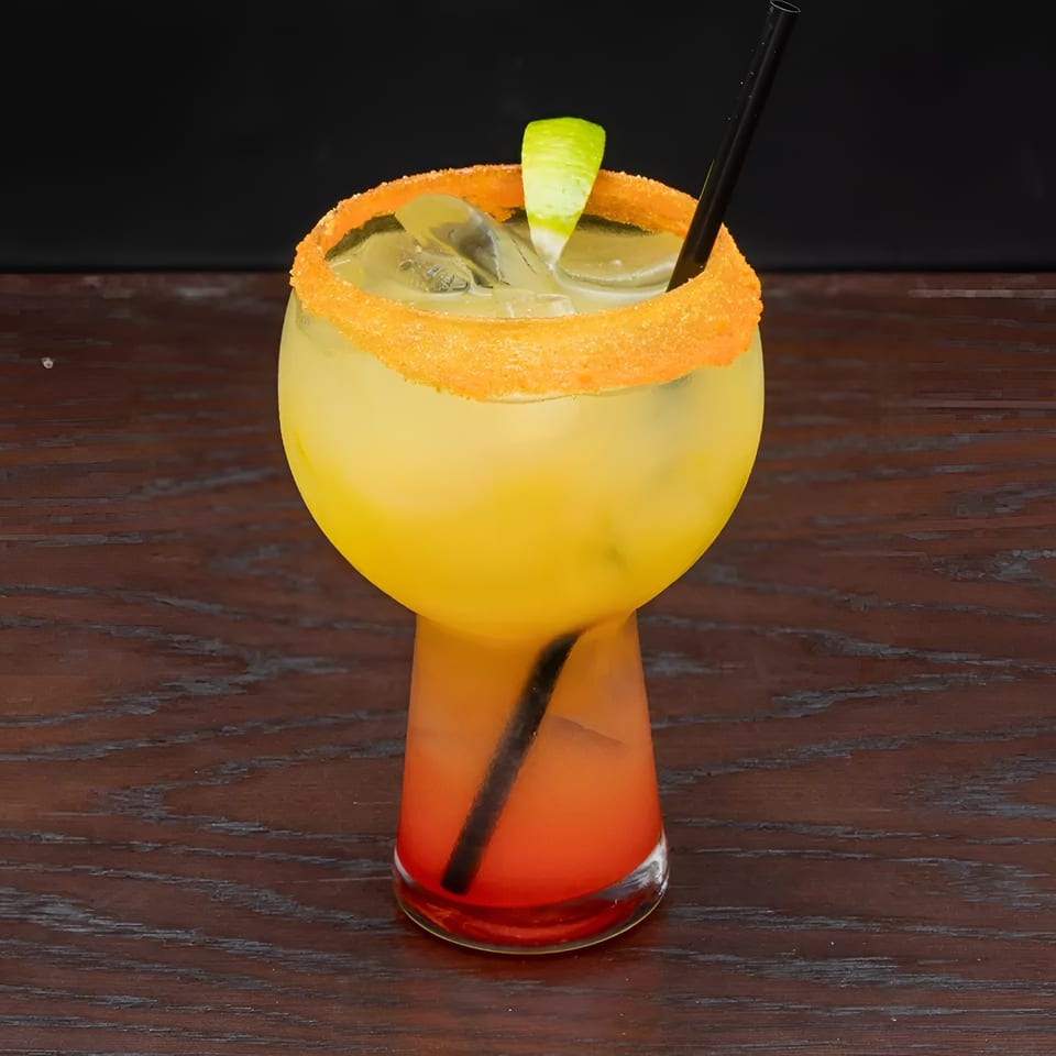 A vibrant yellow and red cocktail with a sugared rim, lime wedge, and straw on a wooden table.
