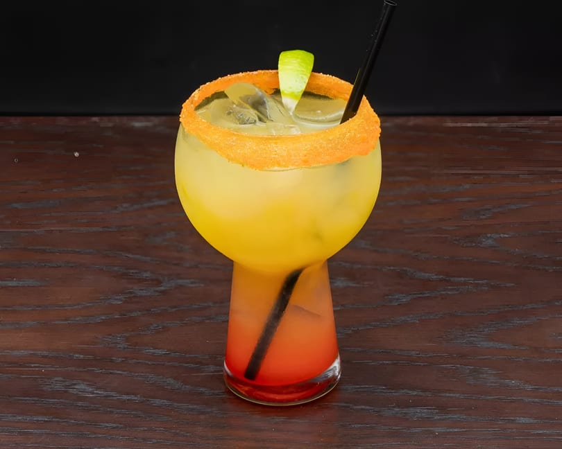 A vibrant yellow and red cocktail with a sugared rim, lime wedge, and straw on a wooden table.