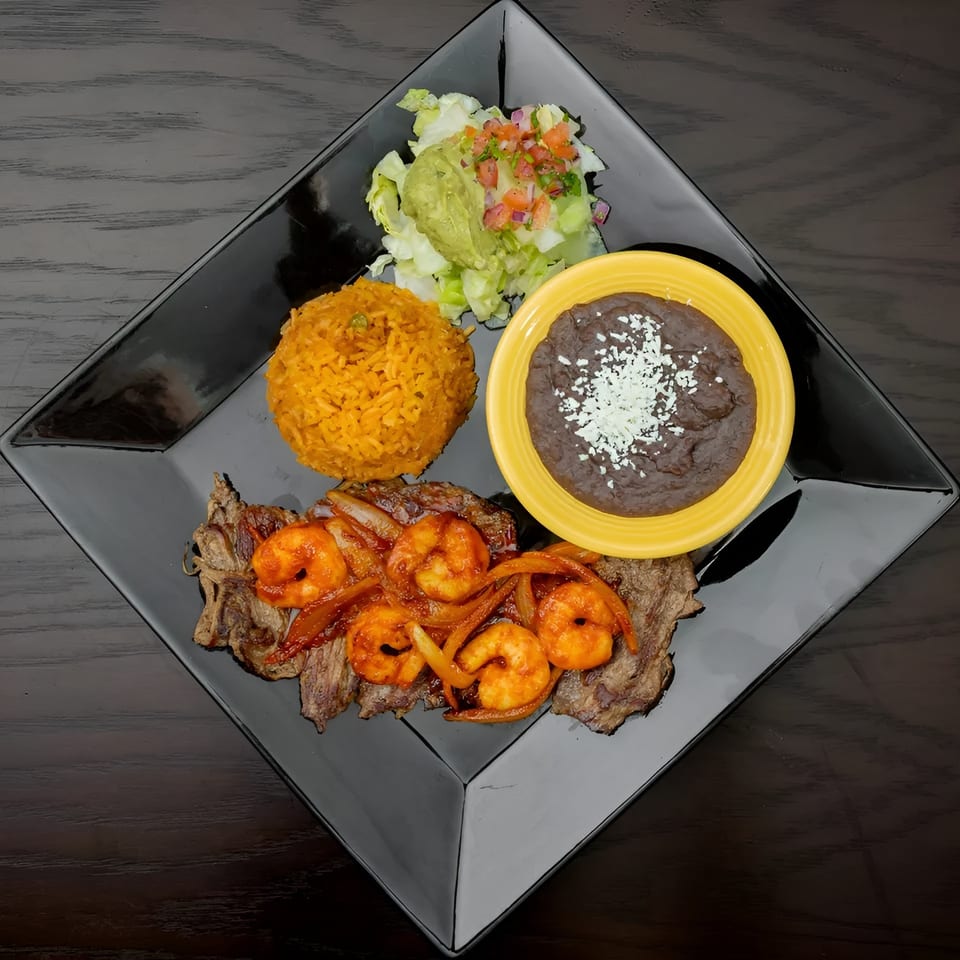 Grilled steak and shrimp with rice, refried beans, guacamole, and pico de gallo on a black plate.
