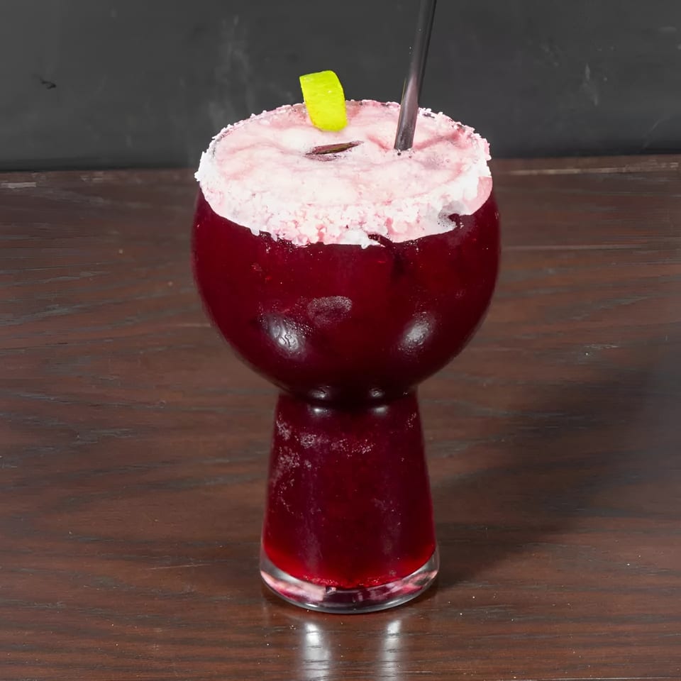 A vibrant red cocktail in a unique glass, topped with pink foam, a lime wedge, and a black straw.