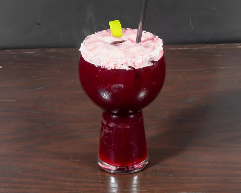 A vibrant red cocktail in a unique glass, topped with pink foam, a lime wedge, and a black straw.