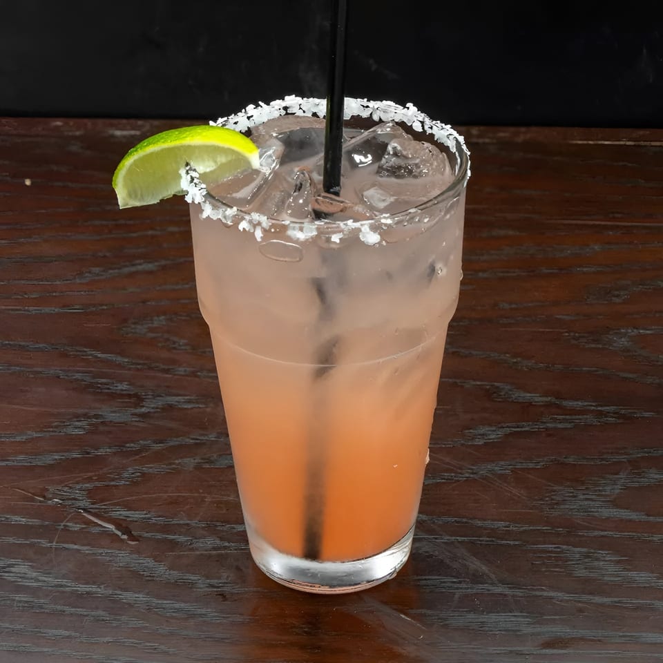 Pink cocktail with ice, a black straw, and a lime wedge, on a wooden table.