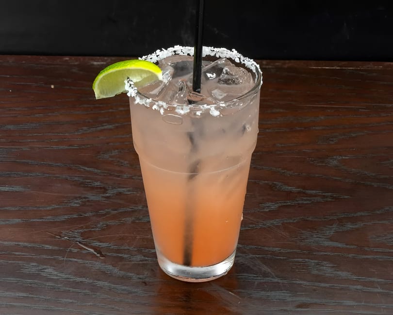 Pink cocktail with ice, a black straw, and a lime wedge, on a wooden table.