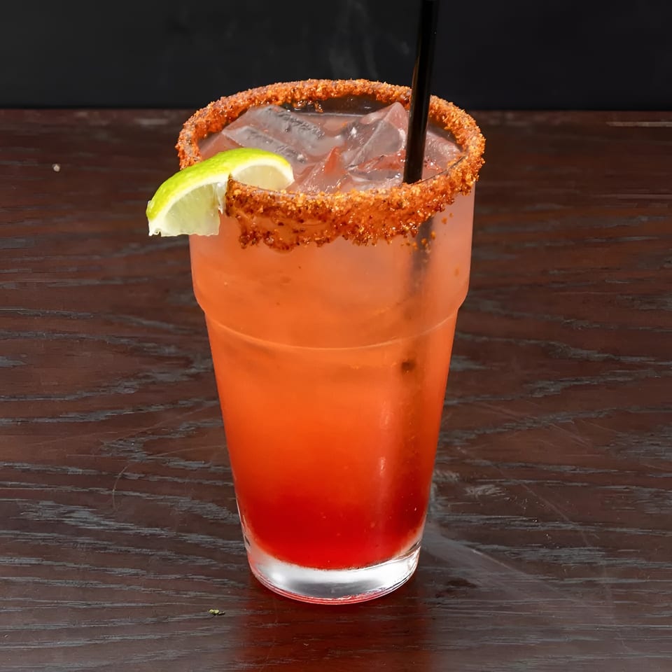 A red cocktail with a chili-lime rim, ice, a lime wedge, and a black straw on a wooden table.
