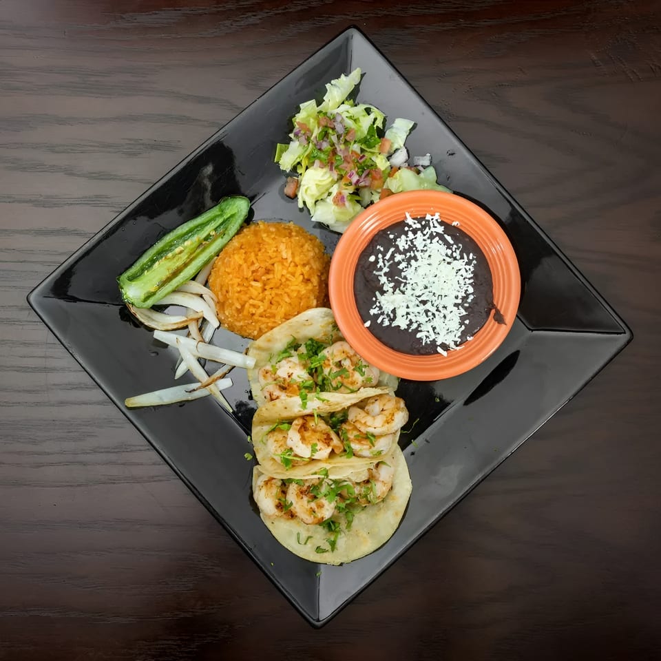 Three shrimp tacos with rice, black beans, salad, grilled jalapeño, and onions on a black plate.