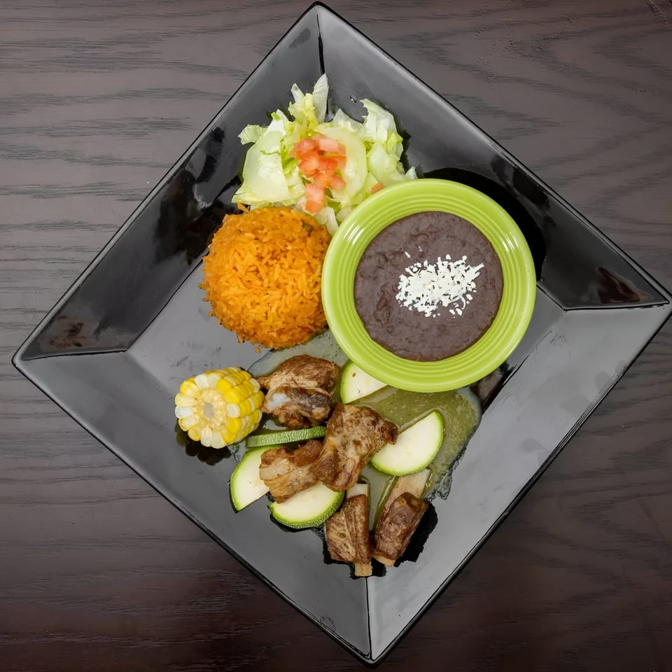 A black square plate with ribs, zucchini, corn, rice, salad, and a bowl of black beans.