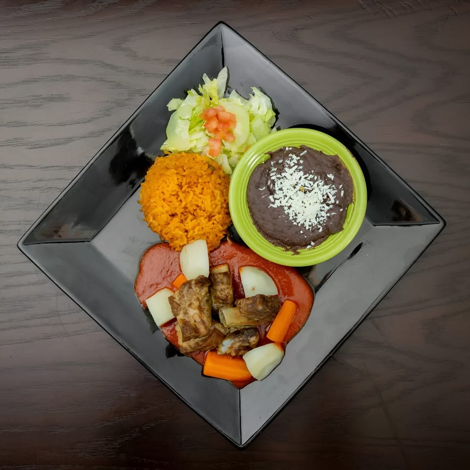 Mexican dish with ribs in red sauce, rice, refried beans, and a side salad on a black plate.