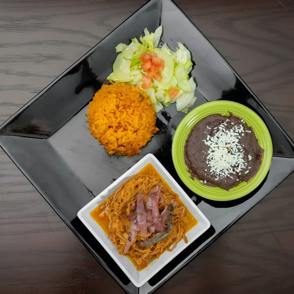 A black square plate holds a Mexican meal with rice, salad, refried beans, and cochinita pibil.