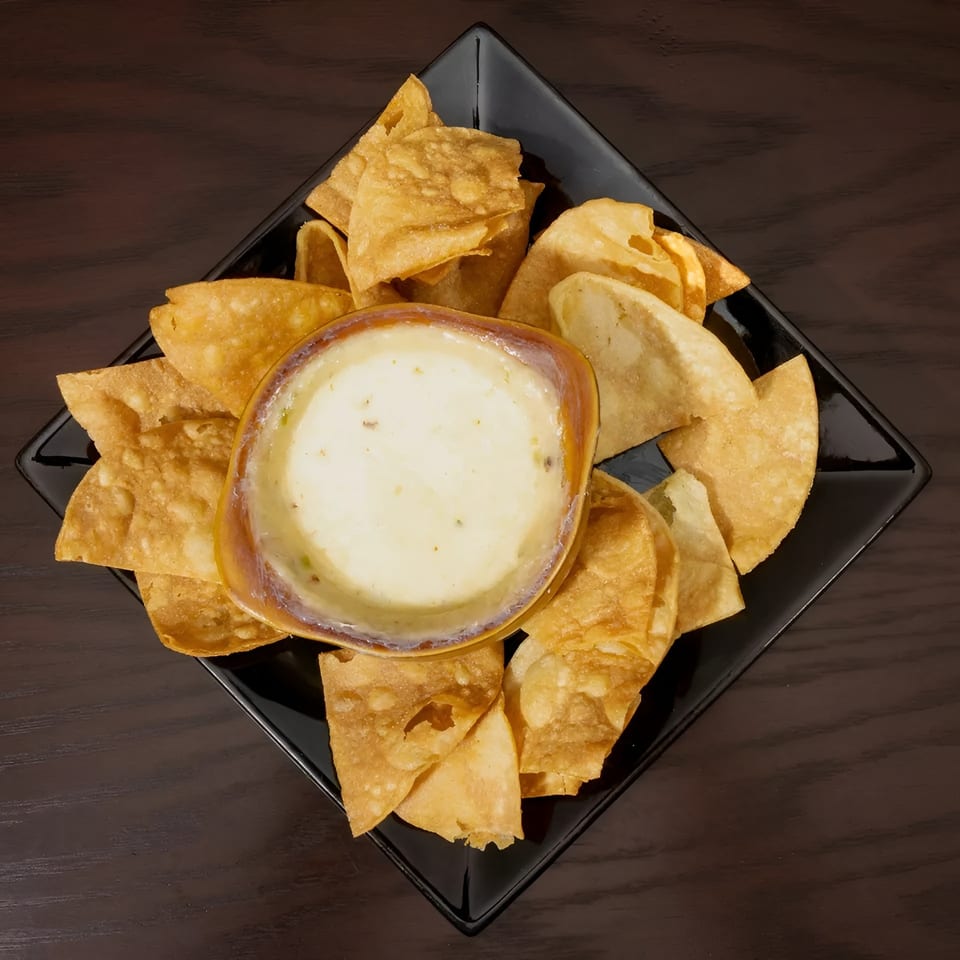 A black square plate holds a bowl of creamy queso dip surrounded by crispy tortilla chips.