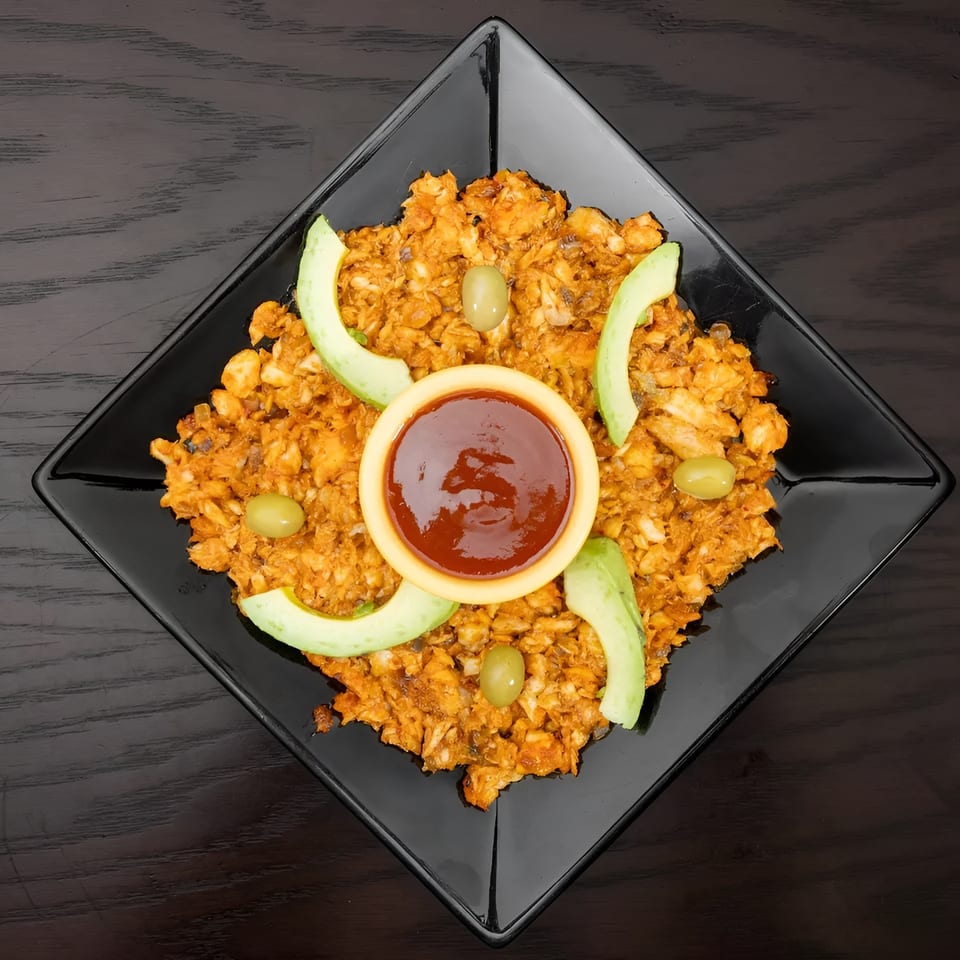 A black square plate holds a mound of orange-brown food, garnished with avocado slices, green olives, and a small bowl of red sauce.