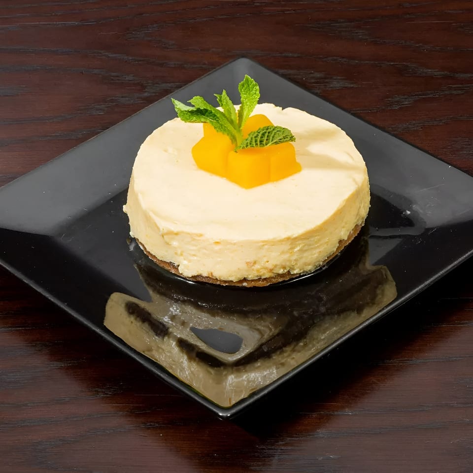 A round cheesecake with mango cubes and mint leaves on a black square plate.